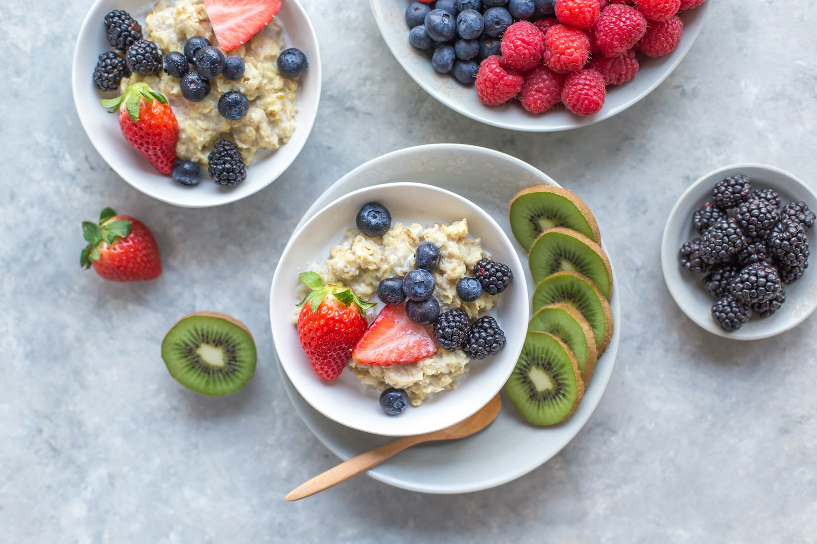 Flat-lay of oatmeal bowls topped with strawberries, blueberries, blackberries and kiwi slices beside bowls of mixed berries on a grey stone surface